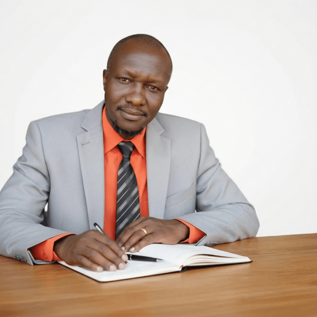 John Monyjok Maluth seated at a desk, focused as he writes in a notebook, symbolizing his dedication to creating life-changing stories that inspire and empower readers.